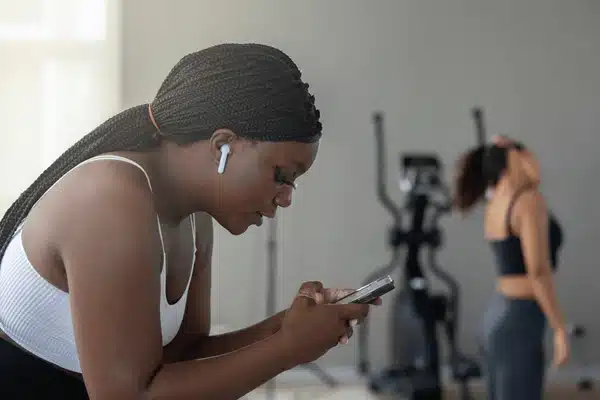 Woman participating in BetterTogether weight loss challenge, using fitness tracking app on smartphone at the gym with friend working out in the background.